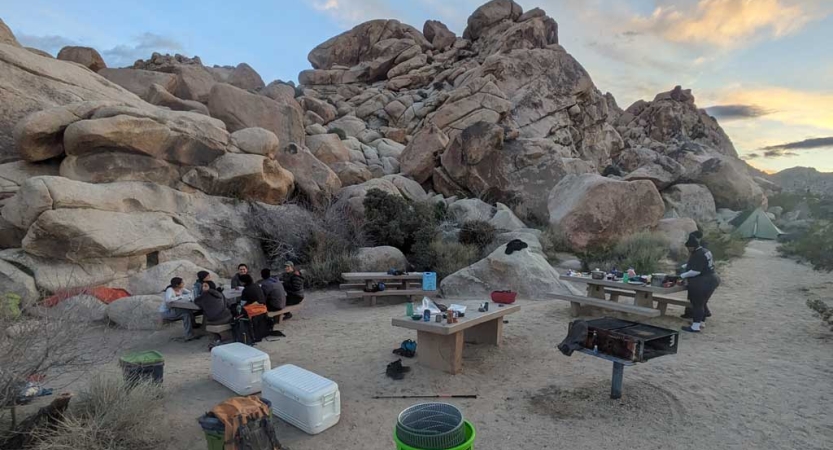 People gather at a campsite near a rock formation in a desert landscape. There is gear scattered on the ground. 